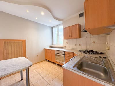 Jasna kuchnia z szafkami z jasnego drewna, podłogą wyłożoną kafelkami, kuchenką gazową, metalowym zlewem, wbudowanym piekarnikiem, dużą ilością miejsca na blacie, małym stolikiem i dużym oknem wpuszczającym naturalne światło - idealna do mieszkania Łódź-Śródmieście na sprzedaż. biuro nieruchomości