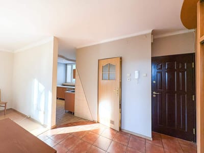 Jasne, puste mieszkanie na sprzedaż w łódzkim Śródmieściu z podłogą wyłożoną kafelkami, beżowymi ścianami, drewnianymi drzwiami, czarnymi drzwiami wejściowymi i częścią kuchenną z drewnianymi szafkami w tle. Przez okno wpada światło słoneczne. biuro nieruchomości