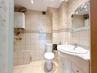 Nowoczesna łazienka w mieszkaniu na sprzedaż w Łodzi-Śródmieściu, z beżowymi kafelkami na ścianach i podłodze, białą toaletą, umywalką z lustrem nad nią, ściennym bojlerem w rogu i wbudowanymi szafkami pod umywalką. biuro nieruchomości