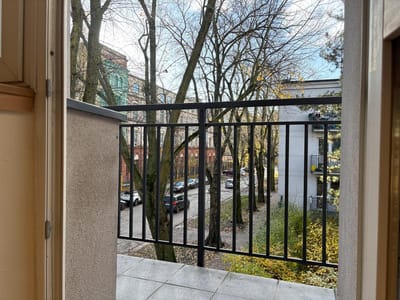 Widok z otwartych drzwi na mały balkon z czarną metalową barierką, wychodzący na ulicę zastawioną zaparkowanymi samochodami i bezlistnymi drzewami w Łodzi-Śródmieściu. Po obu stronach widać budynki - idealne miejsce na mieszkanie na sprzedaż. biuro nieruchomości