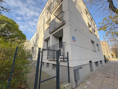 Nowoczesny, jasnoszary apartamentowiec z małymi balkonami i numerem 72 na ścianie, ogrodzony metalowym płotem. Doskonałe mieszkanie na sprzedaż w Śródmieściu, Łódź. Jesienne drzewa i częściowo zachmurzone niebo dopełniają widoku. biuro nieruchomości