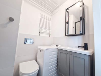 Kompaktowa, nowoczesna łazienka w mieszkaniu na sprzedaż Zduńska Wola posiada białą toaletę obok pralki ładowanej od przodu, szarą toaletkę z czarną baterią, szafkę z lustrem, podgrzewany wieszak na ręczniki i suszarkę sufitową. biuro nieruchomości