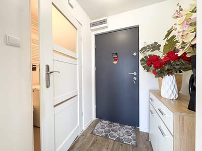 Wejście do mieszkania z ciemnymi drzwiami, dywanikiem i wazonem z kwiatami.