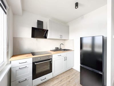 Nowoczesna, kompaktowa kuchnia w mieszkaniu na sprzedaż Zduńska Wola, z jasnoszarymi szafkami, drewnianym blatem, czarną kuchenką elektryczną, piekarnikiem, zlewem i matową czarną lodówką. Nad kuchenką wisi okap, a przez okno wpada naturalne światło. biuro nieruchomości