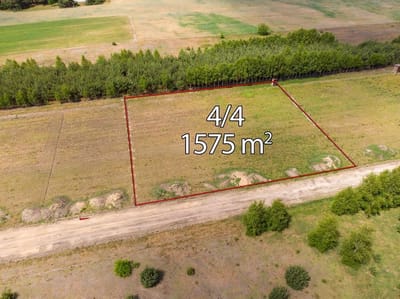 Widok z lotu ptaka na prostokątną działkę zaznaczoną na czerwono, oznaczoną numerem 4/4 o powierzchni 1575 m². Działka na sprzedaż Skotniki - łódzkie znajduje się przy drodze gruntowej, graniczy z drzewami i otoczona jest otwartymi terenami trawiastymi. biuro nieruchomości