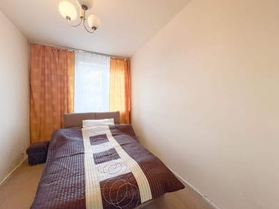 Mała, jasna sypialnia w mieszkaniu na sprzedaż w Łodzi-Retkini posiada podwójne łóżko z brązowo-beżową kołdrą, jasne ściany, górne oświetlenie, pomarańczowe zasłony i okno wpuszczające naturalne światło. biuro nieruchomości