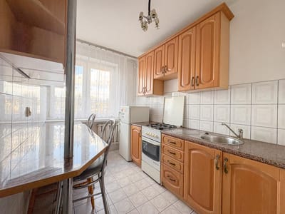 Przytulna kuchnia w Łodzi-Retkini wyposażona jest w szafki z jasnego drewna, białą kuchenkę gazową, kuchenkę mikrofalową i stół jadalny przy dużym oknie z zasłonami. Podłoga wyłożona kafelkami dodaje uroku temu przytulnemu mieszkaniu na sprzedaż. biuro nieruchomości