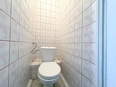 Mała łazienka w mieszkaniu na sprzedaż, z białą toaletą na tle ścian pokrytych białymi płytkami z czerwoną fugą. Pomieszczenie jest wąskie i jasno oświetlone z góry, oferując czystą i nowoczesną przestrzeń w Łodzi-Retkini. biuro nieruchomości