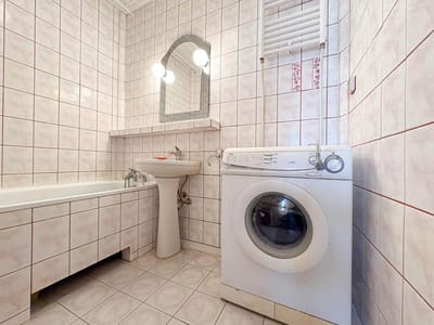 Mała łazienka w mieszkaniu na sprzedaż w Łodzi-Retkini, z różowawymi kafelkami, wanną, białą umywalką z lustrzanym oświetleniem nad nią i pralką ładowaną od przodu obok podgrzewanego wieszaka na ręczniki. biuro nieruchomości