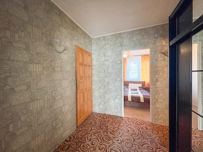 Przedpokój z wzorzystą tapetą i wykładziną, drewniane drzwi po lewej stronie i otwarte drzwi prowadzące do sypialni z dużym oknem i pomarańczowymi zasłonami - idealne dla osób poszukujących mieszkania na sprzedaż w Łodzi-Retkini. biuro nieruchomości