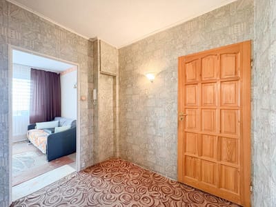 Przedpokój z wzorzystą tapetą i dywanem, drewniane drzwi po prawej stronie, lampa wisząca na ścianie i widok na salon z sofą i oknem z zasłonami w tym mieszkaniu na sprzedaż w Łodzi-Retkini. biuro nieruchomości