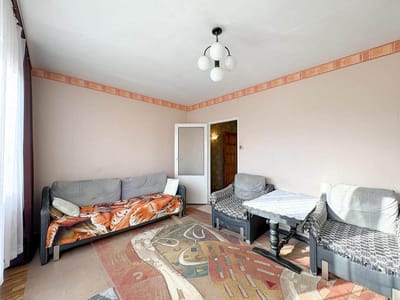 Salon w Łodzi-Retkini z beżowymi ścianami, dwoma szarymi fotelami, szarą sofą z kocem w tygrysie wzory, wzorzystym dywanem, białymi drzwiami i lampą sufitową - idealne mieszkanie na sprzedaż. biuro nieruchomości