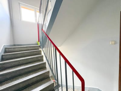 Jasno oświetlone schody wewnętrzne z szarymi stopniami i czerwoną poręczą prowadzą na podest ze światłem słonecznym wpadającym przez okno - idealne do mieszkania Łódź-Retkinia na sprzedaż. Białe ściany i włącznik światła po prawej stronie dodają nowoczesności. biuro nieruchomości
