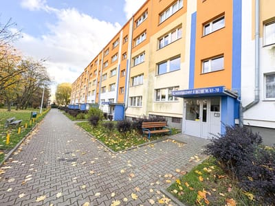 Długi, czteropiętrowy apartamentowiec z pomarańczowo-żółtymi ścianami i niebieskimi akcentami oferuje mieszkanie na sprzedaż w Łodzi-Retkini. Opadłe liście pokrywają wybrukowany chodnik, otoczony ławkami i krzewami pod częściowo zachmurzonym niebem. biuro nieruchomości