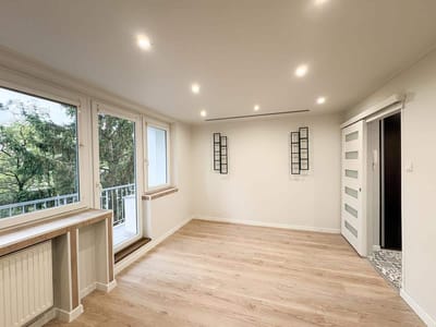 Wnętrze mieszkania z drewnianą podłogą, dużymi oknami i nowoczesnymi elementami wykończenia.