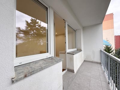 Długi, wąski balkon z szarą podłogą z płytek i metalową balustradą wychodzi na drzewa. Dwa duże okna i otwarte drzwi prowadzą do 2 pokojowego mieszkania z beżową tapetą w Łodzi-Retkini. W pobliżu widoczna jest biała elewacja i kolorowy budynek. biuro nieruchomości