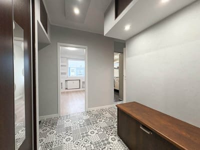 Nowoczesny przedpokój w tym mieszkaniu w Łodzi-Retkini ma wzorzystą podłogę z płytek, jasnoszare ściany, wbudowane lampy sufitowe, drewnianą ławkę ze schowkiem i wykończenia z ciemnego drewna. Korytarz prowadzi do jasnego pokoju z drewnianą podłogą i białymi szafkami. biuro nieruchomości