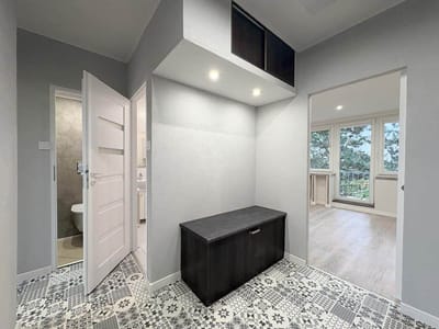 Nowoczesny przedpokój w tym mieszkaniu w Łodzi-Retkini ma wzorzystą podłogę z płytek, czarną ławkę do przechowywania i górne szafki. Po lewej stronie znajdują się drzwi do łazienki, a po prawej otwarte drzwi prowadzą do jasnego 2-pokojowego pomieszczenia z dużymi oknami. biuro nieruchomości