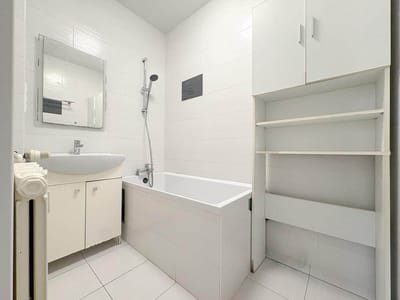 Biała, minimalistyczna łazienka w mieszkaniu w Łodzi-Retkini obejmuje wannę z zamontowanym na ścianie prysznicem, umywalkę i szafkę, lustro nad umywalką oraz otwarte półki. Całkowicie białe płytki dodają czystego, jasnego charakteru w tym 2 pokojowym mieszkaniu. biuro nieruchomości