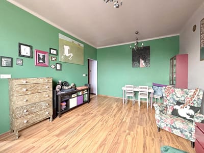 Jasny salon-jadalnia z zielonymi ścianami, drewnianą podłogą, fotelem w kwiaty, wzorzystą skrzynią, grafiką na ścianie, półkami do przechowywania, stołem z czterema krzesłami i dwoma żyrandolami. biuro nieruchomości