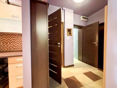 Korytarz z jasnofioletowymi ścianami, podłogą wyłożoną kafelkami i dwojgiem ciemnobrązowych drzwi, z których jedne są otwarte, a drugie uchylone. Po lewej stronie widać część biurka i mozaikowy backsplash. biuro nieruchomości