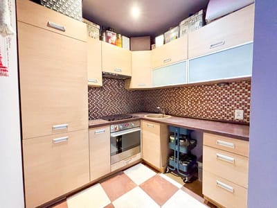 Nowoczesna kuchnia z szafkami z jasnego drewna, wbudowanym piekarnikiem, płytą elektryczną, małym okrągłym zlewem, mozaikowym backsplash i brązowo-białymi płytkami podłogowymi w kratkę. Blaty są ciemne, a na półkach znajdują się różne przedmioty kuchenne. biuro nieruchomości