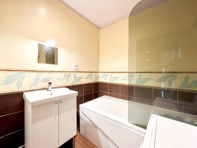 Mała łazienka z białą szafką pod umywalkę, prostokątną wanną, lustrem ściennym nad umywalką i dekoracyjnymi płytkami z liściastym wzorem na ścianie. biuro nieruchomości