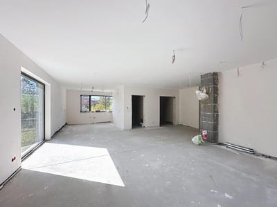 Puste, niewykończone pomieszczenie z białymi ścianami, dużymi oknami po lewej stronie i odsłoniętym okablowaniem zwisającym z sufitu w domu na sprzedaż Radomsko - Wygoda. W pobliżu znajdują się dwa otwarte otwory drzwiowe i filar z materiałami budowlanymi. biuro nieruchomości