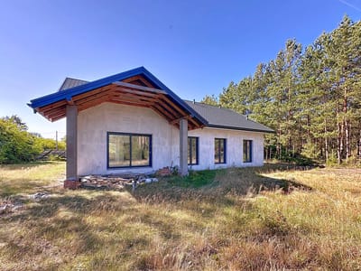 Częściowo zbudowany, jednopiętrowy dom na sprzedaż Radomsko - Wygoda ma ciemny dach i niedokończony wygląd zewnętrzny, ustawiony na trawiastej działce obok wysokich sosen pod czystym, błękitnym niebem. biuro nieruchomości