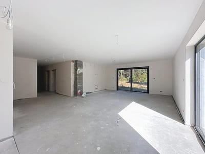 Przestronny, niewykończony pokój z gołą betonową podłogą, białymi ścianami, odsłoniętym okablowaniem i dużymi szklanymi drzwiami wpuszczającymi naturalne światło, z widokiem na zieleń - idealny dla osób poszukujących domu na sprzedaż Radomsko - Wygoda. biuro nieruchomości