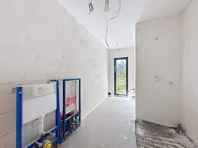 Pokój w trakcie remontu w domu na sprzedaż w Radomsku, z odsłoniętymi mediami, niedokończonymi ścianami i gołą podłogą. Z sufitu zwisają przewody elektryczne, a duże okno na drugim końcu wypełnia przestrzeń naturalnym światłem. biuro nieruchomości