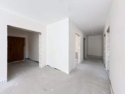 Korytarz w budynku w budowie z niedokończonymi białymi ścianami, odsłoniętym okablowaniem i betonową podłogą - idealny dla tych, którzy szukają domu na sprzedaż Radomsko. Drewniane drzwi stoją na końcu jednego otwartego wejścia, zapraszając wzrok. biuro nieruchomości