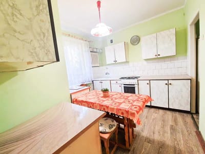 Mała kuchnia z zielonymi ścianami, białymi szafkami, kuchenką i zegarem. Na środku stoi drewniany stół przykryty czerwonym wzorzystym obrusem i dwa krzesła. Światło wpada przez okno z przezroczystymi zasłonami. biuro nieruchomości