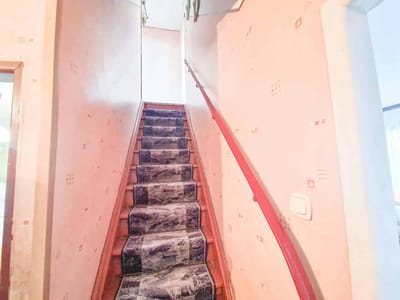 Wąskie schody z wzorzystym dywanem i czerwoną poręczą wznoszą się między dwiema bladoróżowymi, wzorzystymi ścianami w dobrze oświetlonym wnętrzu. biuro nieruchomości