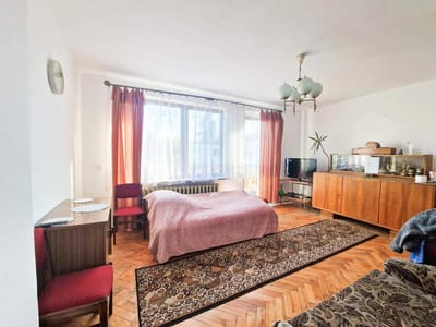 Jasna sypialnia w domu na sprzedaż w Pabianicach z dużym oknem, przezroczystymi i rdzawymi zasłonami, różowym łóżkiem, czerwonymi krzesłami, wzorzystym dywanem, sofą, drewnianą szafką ze szklanymi naczyniami, telewizorem i rośliną doniczkową. biuro nieruchomości