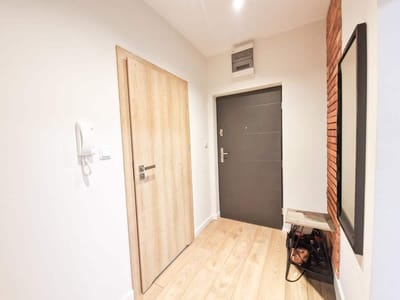 Nowoczesny, minimalistyczny przedpokój w mieszkaniu na sprzedaż Łódź Śródmieście Tuwima z podłogą i drzwiami z jasnego drewna, ciemnoszarymi drzwiami wejściowymi, telefonem zamontowanym na ścianie, dużym lustrem, małą półką nad torbą i butami oraz industrialną ścianą z czerwonej cegły. biuro nieruchomości