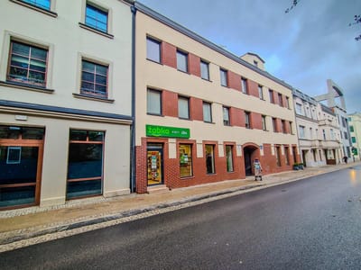 Spokojna miejska ulica z nowoczesnymi, trzypiętrowymi budynkami. Mały zielony szyld sklepu Żabka oznacza sklep w pobliżu mieszkania na sprzedaż Łódź Śródmieście Tuwima, a jedna osoba idzie pustym, spokojnym chodnikiem pod zachmurzonym niebem. biuro nieruchomości