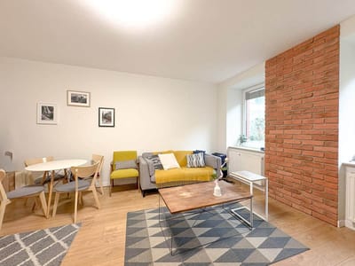 Jasny salon w mieszkaniu na sprzedaż Łódź Śródmieście Tuwima ma drewniane podłogi, szaro-żółtą sofę, mały biały stolik kawowy, jadalnię z okrągłym stołem, ceglaną ścianę akcentującą, okno, oprawioną sztukę i wzorzysty dywan. biuro nieruchomości