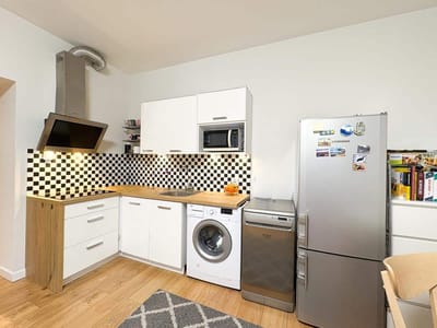 Nowoczesna kuchnia w mieszkaniu na sprzedaż Łódź Śródmieście Tuwima - białe szafki, drewniany blat, backsplash w czarno-białą kratkę, okap ze stali nierdzewnej, pralka, zmywarka, srebrna lodówka z magnesami. Półka na kubki, widoczne książki kucharskie i owoce. biuro nieruchomości