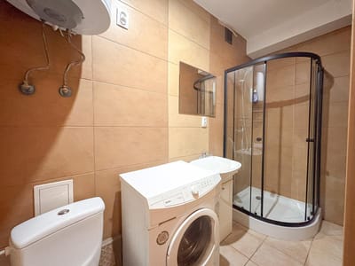 Niewielka łazienka w kawalerce na sprzedaż w Łodzi-Bałutach ma ściany wyłożone brązowymi kafelkami, toaletę, białą pralkę, umywalkę z lustrem, narożną szklaną kabinę prysznicową oraz widoczne rury i podgrzewacz wody nad toaletą. biuro nieruchomości