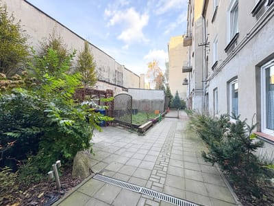 Wybrukowane podwórko wzdłuż jasnego budynku w Łodzi-Bałutach, z zielonymi krzewami i małymi drzewami po obu stronach, ogrodzony ogródek przed nim i częściowo zachmurzone błękitne niebo nad nim - idealne na nową kawalerkę na sprzedaż. biuro nieruchomości