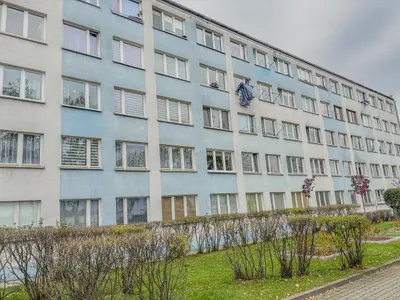 Budynek mieszkalny z wieloma oknami, niektóre z zasłonami, inne otwarte, z widocznymi roślinami przed budynkiem.