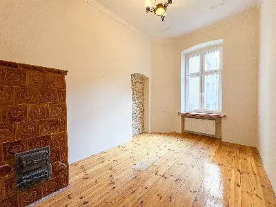 Salon z kaflowym piecem, drewnianą podłogą i wysokim oknem w kamienicy