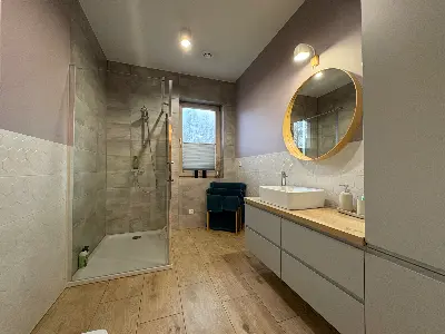 Duża łazienka z kabiną prysznicową, okrągłym lustrem w złotej ramie i szafką podumywalkową