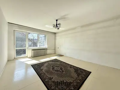 Pusty pokój z kafelkowaną podłogą, dywanem i grzejnikiem pod oknem — do remontu