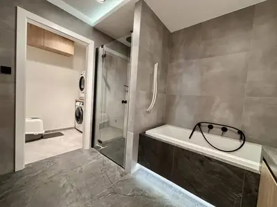 Łazienka z wanną i czarną armaturą, prysznicem za szybą, szarymi płytkami i wejściem do pralni