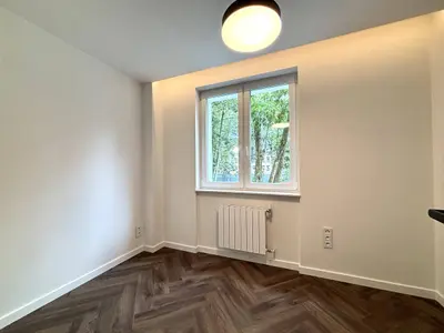 Mała sypialnia z białymi ścianami, parkietem i oknem wychodzącym na drzewa