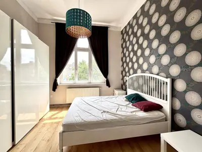 Sypialnia z białym łóżkiem, tapetą w koła, zasłonami bordowymi i turkusową lampą