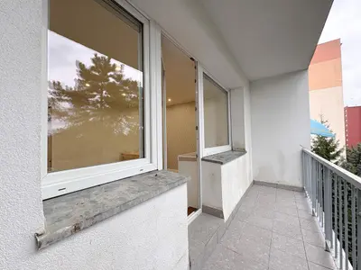 Balkon z szarą podłogą z płytek i metalową balustradą, widok na drzewa i okoliczne bloki.