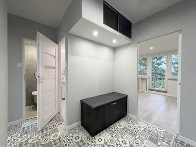 Przedpokój z wzorzystą podłogą i czarną ławką, otwarte przejście do salonu z dużymi oknami.